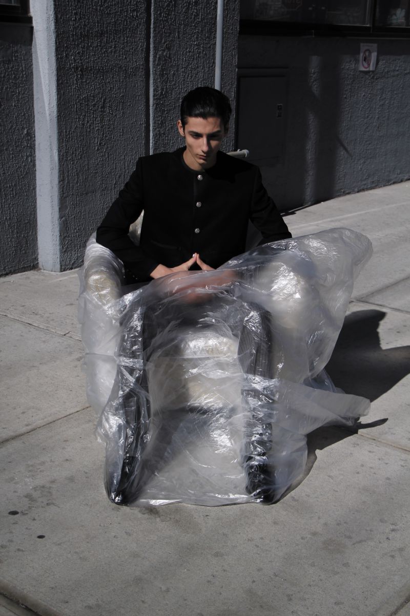 Stylish man in black jacket sits in an armchair wrapped in plastic, outdoors against a grey building. Shadow and sunlight.