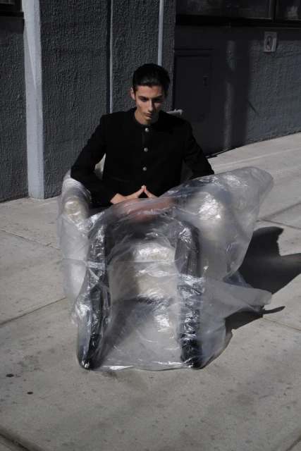 Stylish man in black jacket sits in an armchair wrapped in plastic, outdoors against a grey building. Shadow and sunlight.
