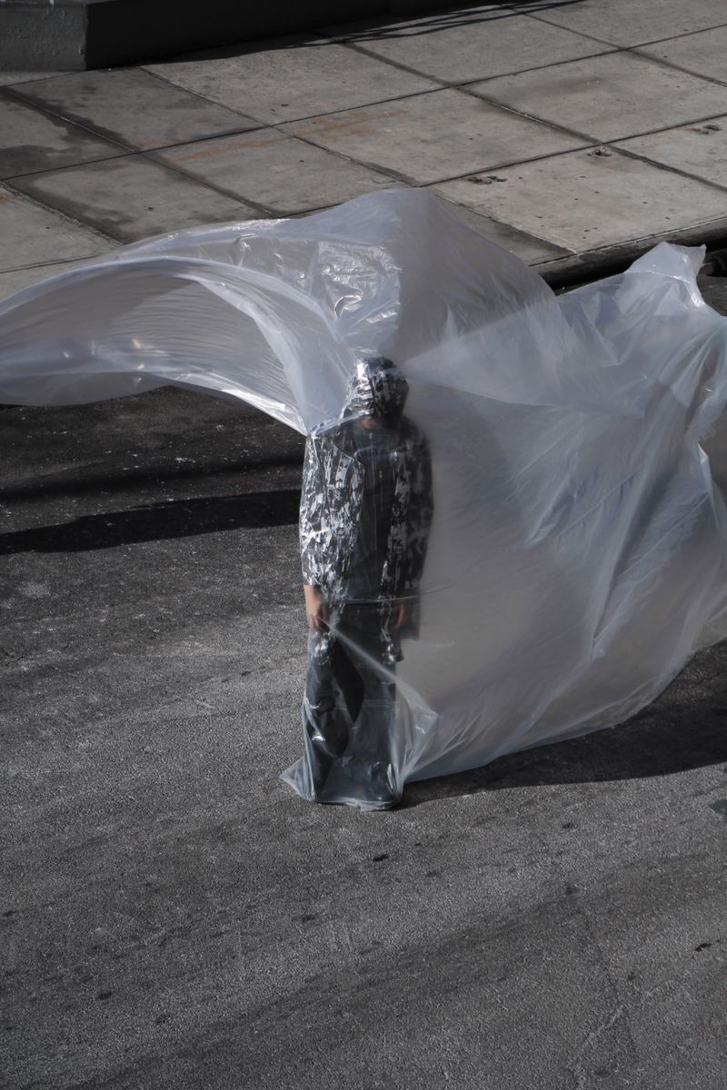 Person wearing a metallic mask and jacket, standing on an asphalt road, partially covered by a sheet of clear plastic.