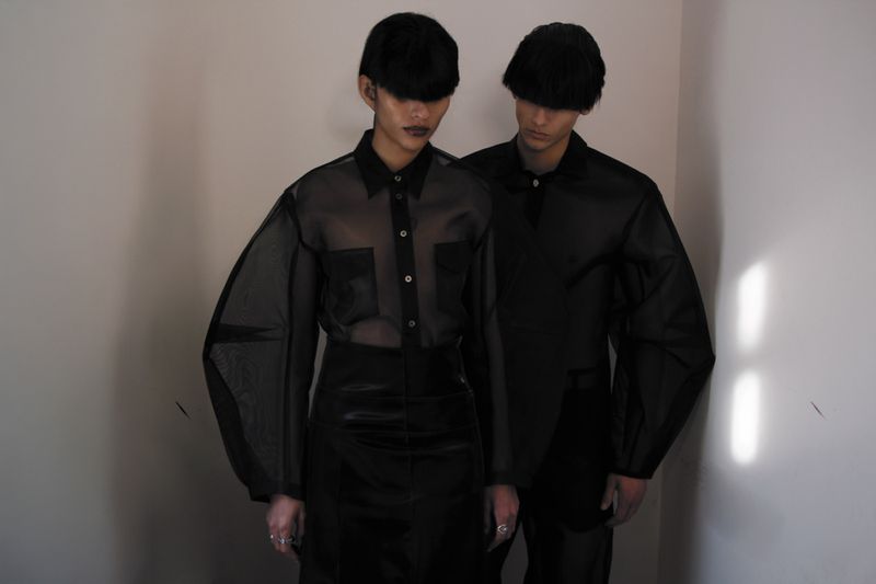Two fashion models in semi-sheer black button-down shirts, black hats and skirts, standing in a corner with minimalist studio lighting.