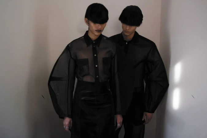 Two fashion models in semi-sheer black button-down shirts, black hats and skirts, standing in a corner with minimalist studio lighting.