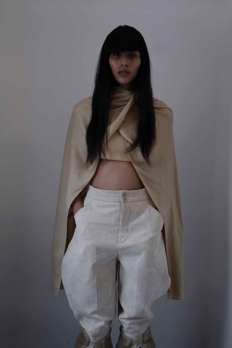A woman with long dark hair stands in a photo studio, wearing a tan draped top and light linen pants with interesting leg shape.