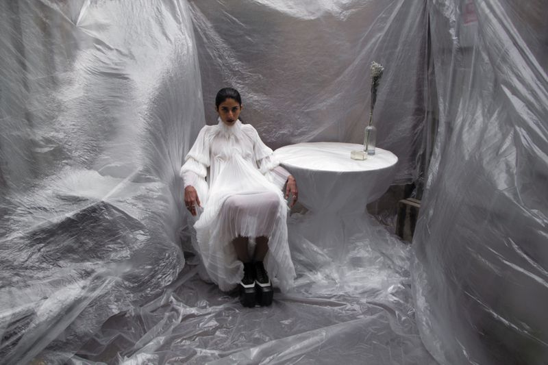 Woman in a white dress sits in a clear-plastic-draped room. A small white table holds a vase. The scene has an editorial feel.