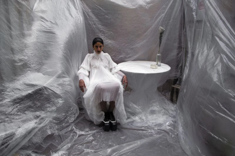 Woman in a white dress sits in a clear-plastic-draped room. A small white table holds a vase. The scene has an editorial feel.