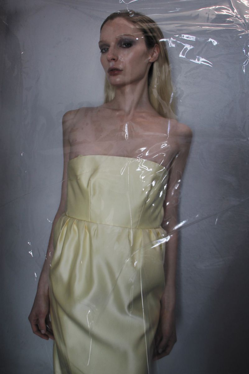 Woman in a light yellow strapless dress stands indoors, partially obscured by a sheet of clear plastic creating an ethereal effect.