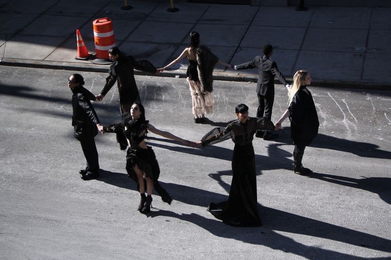 Seven fashion models, diverse genders and ethnicities, form a circle on an urban street, wearing dark and eclectic clothing.