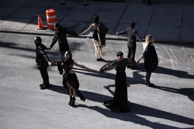Seven fashion models, diverse genders and ethnicities, form a circle on an urban street, wearing dark and eclectic clothing.