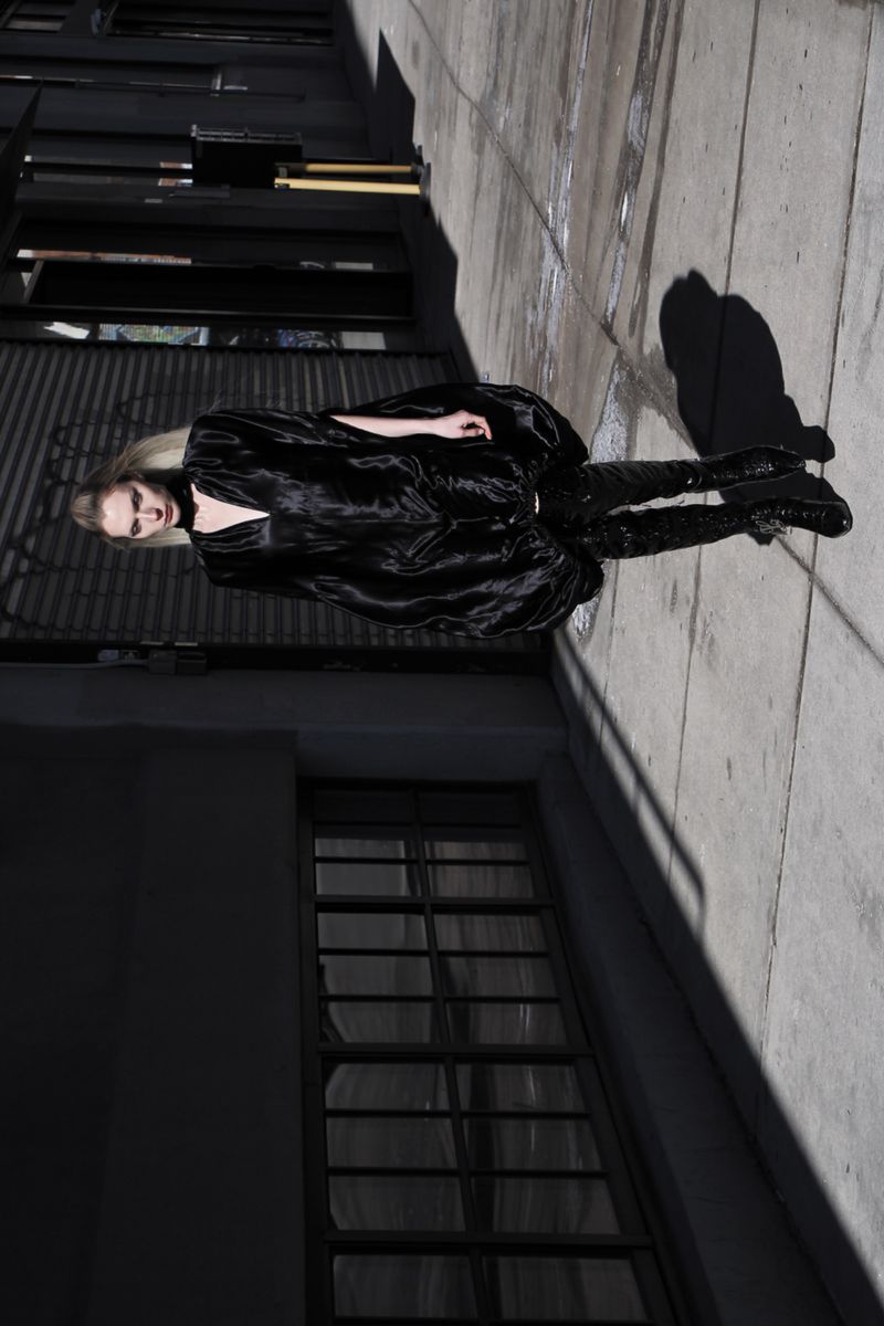 A fashionably dressed woman in a black outfit and boots poses in an alleyway bathed in sunlight, casting long shadows on the pavement.