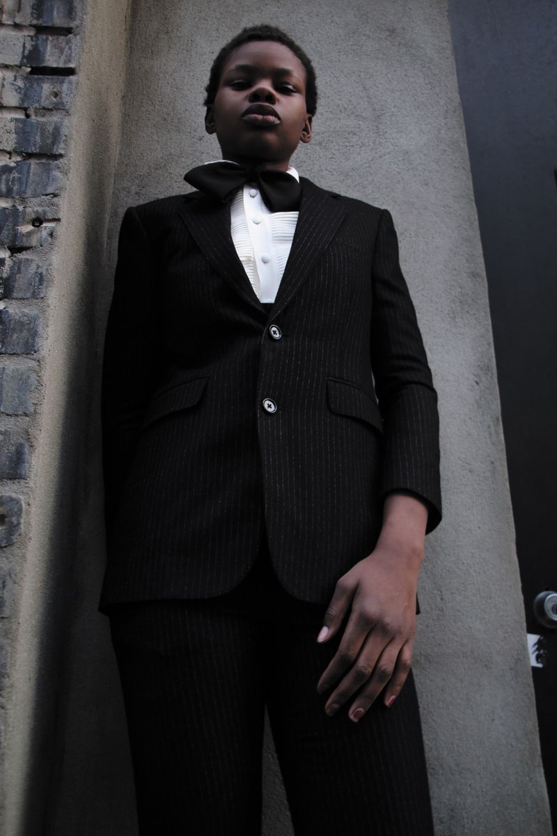 Person in a suit and bow tie stands against a textured wall. The ensemble includes a black suit, crisp white shirt, and black bow tie.
