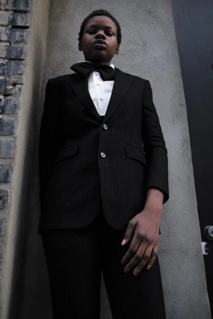 Person in a suit and bow tie stands against a textured wall. The ensemble includes a black suit, crisp white shirt, and black bow tie.