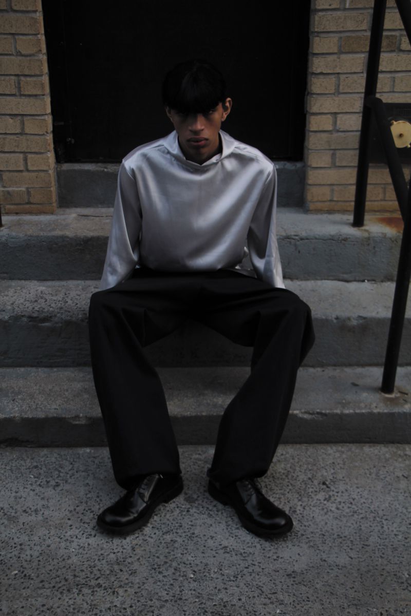 Adult male in a silver top and black trousers poses on concrete steps in front of a dark doorway flanked by brick walls.