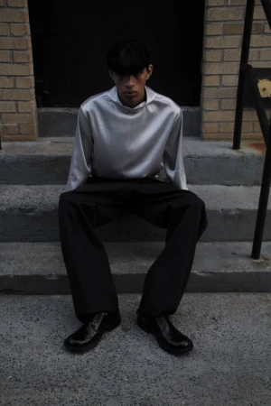 Adult male in a silver top and black trousers poses on concrete steps in front of a dark doorway flanked by brick walls.