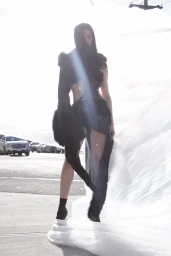 Model standing outdoors next to a draped white fabric with parked cars visible in the background.