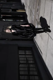 Model wearing a voluminous black dress and tall boots standing on a concrete sidewalk in front of a dark building exterior.