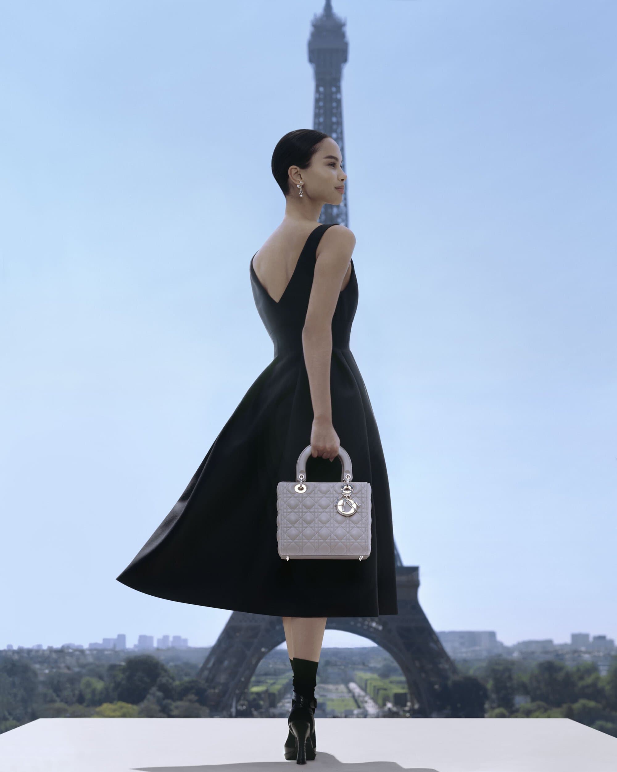 Elegant woman in a black dress poses with a handbag in front of the Eiffel Tower in Paris, evoking classic fashion and style.