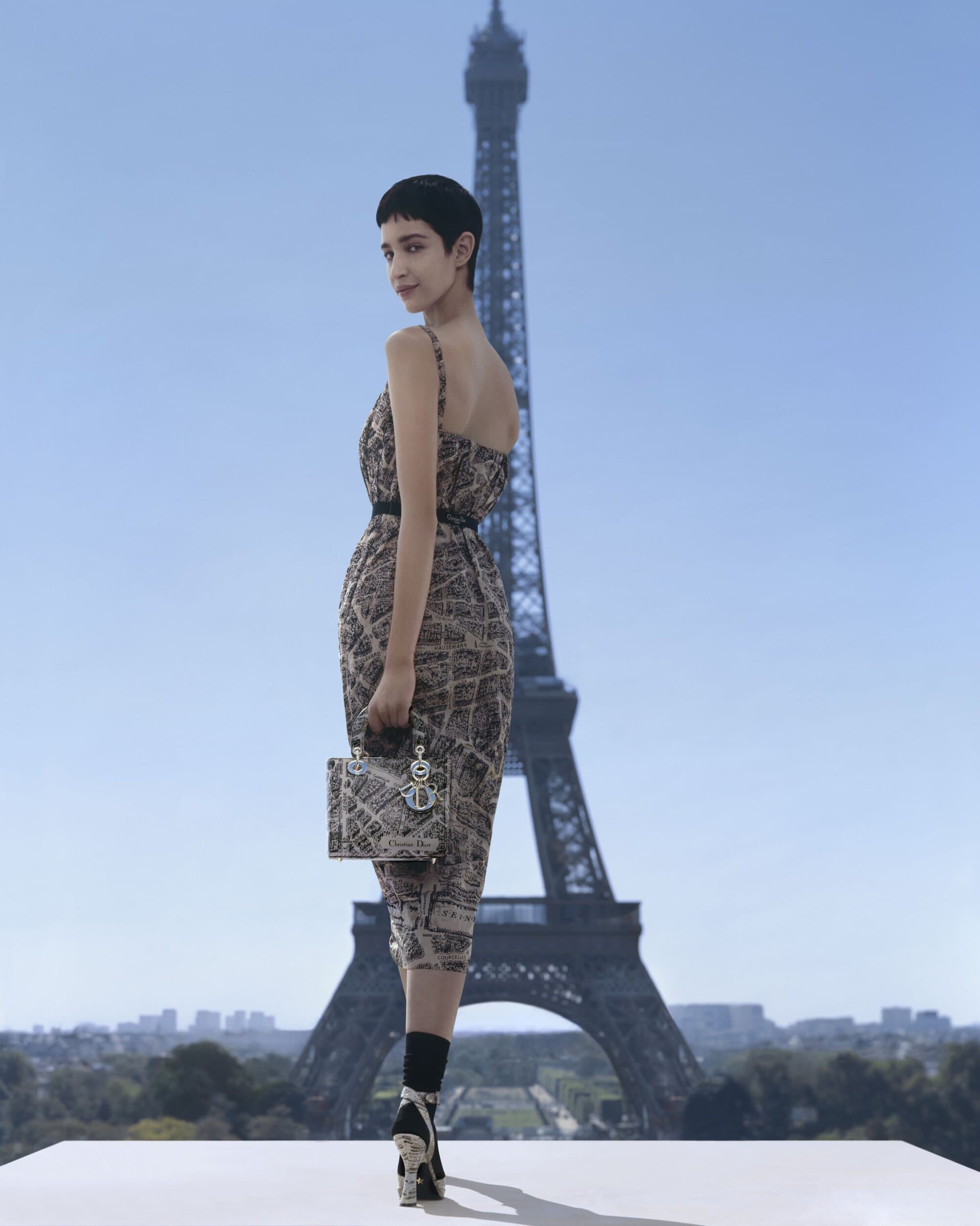 Model in a map-printed dress holding a handbag with the Eiffel Tower centered in the background under a clear sky.
