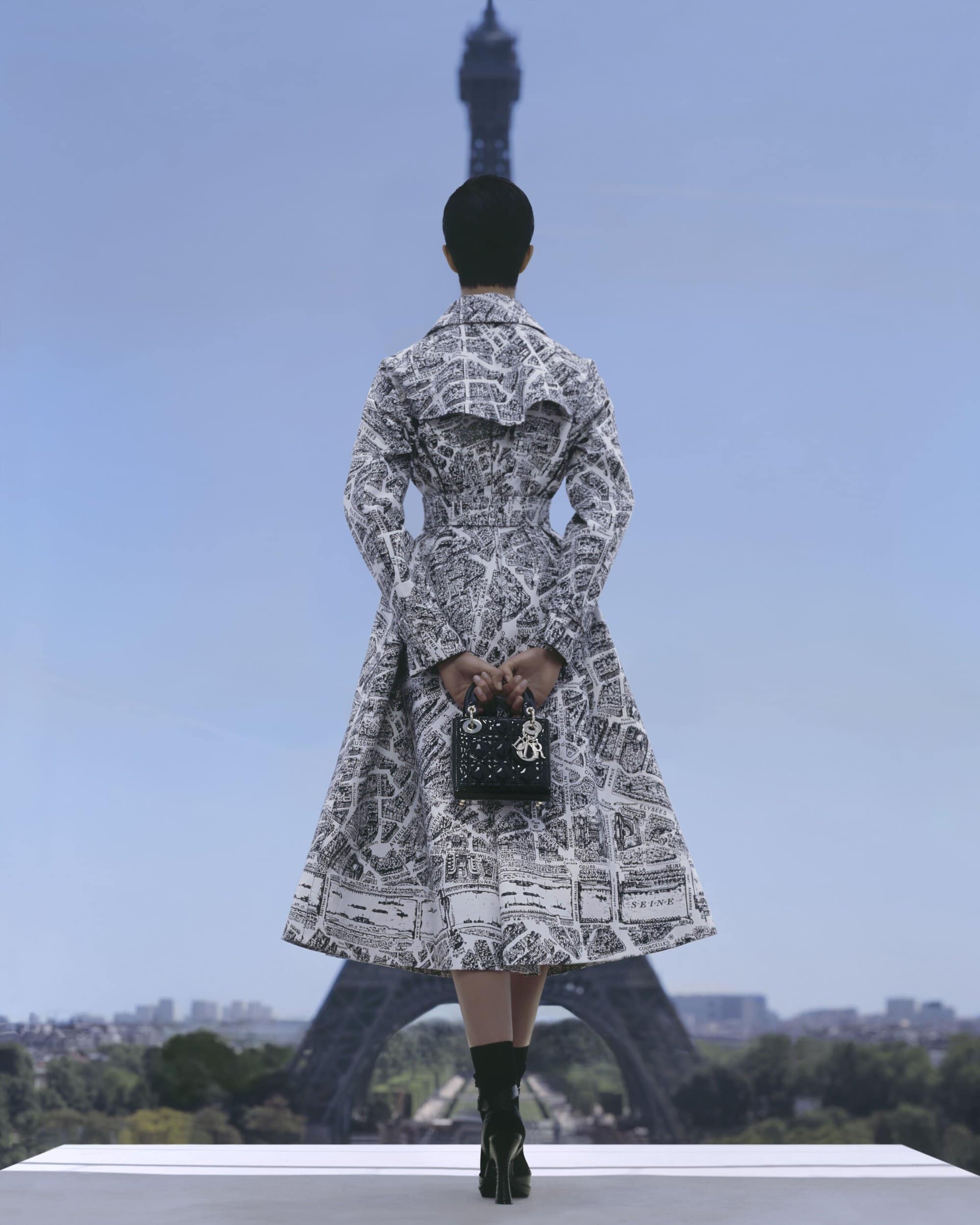 Woman in a printed dress and boots stands on a white platform with the Eiffel Tower in the background on a clear day.