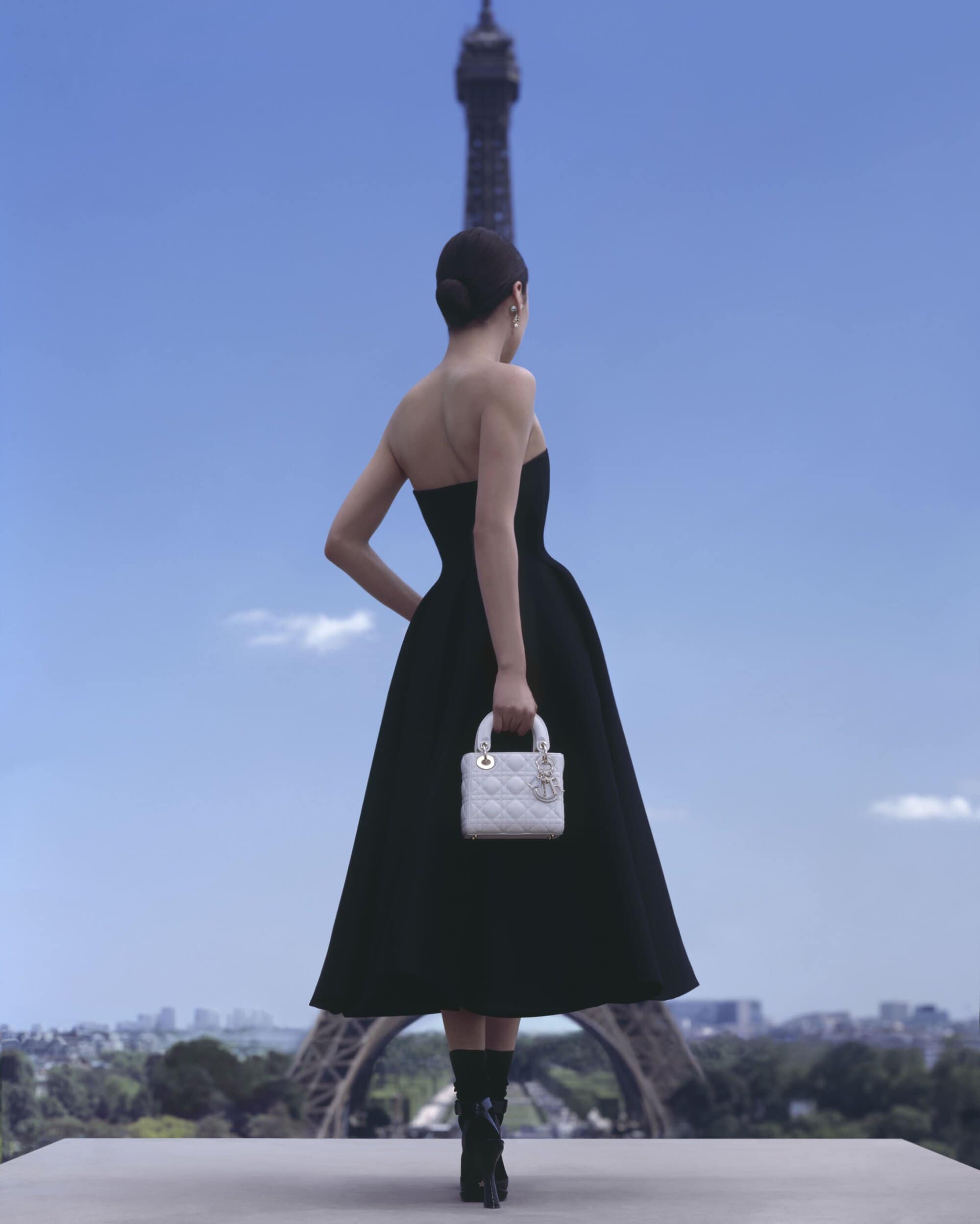 Woman in a black dress holding a handbag stands against a backdrop of the Eiffel Tower and the Paris skyline under a bright blue sky.