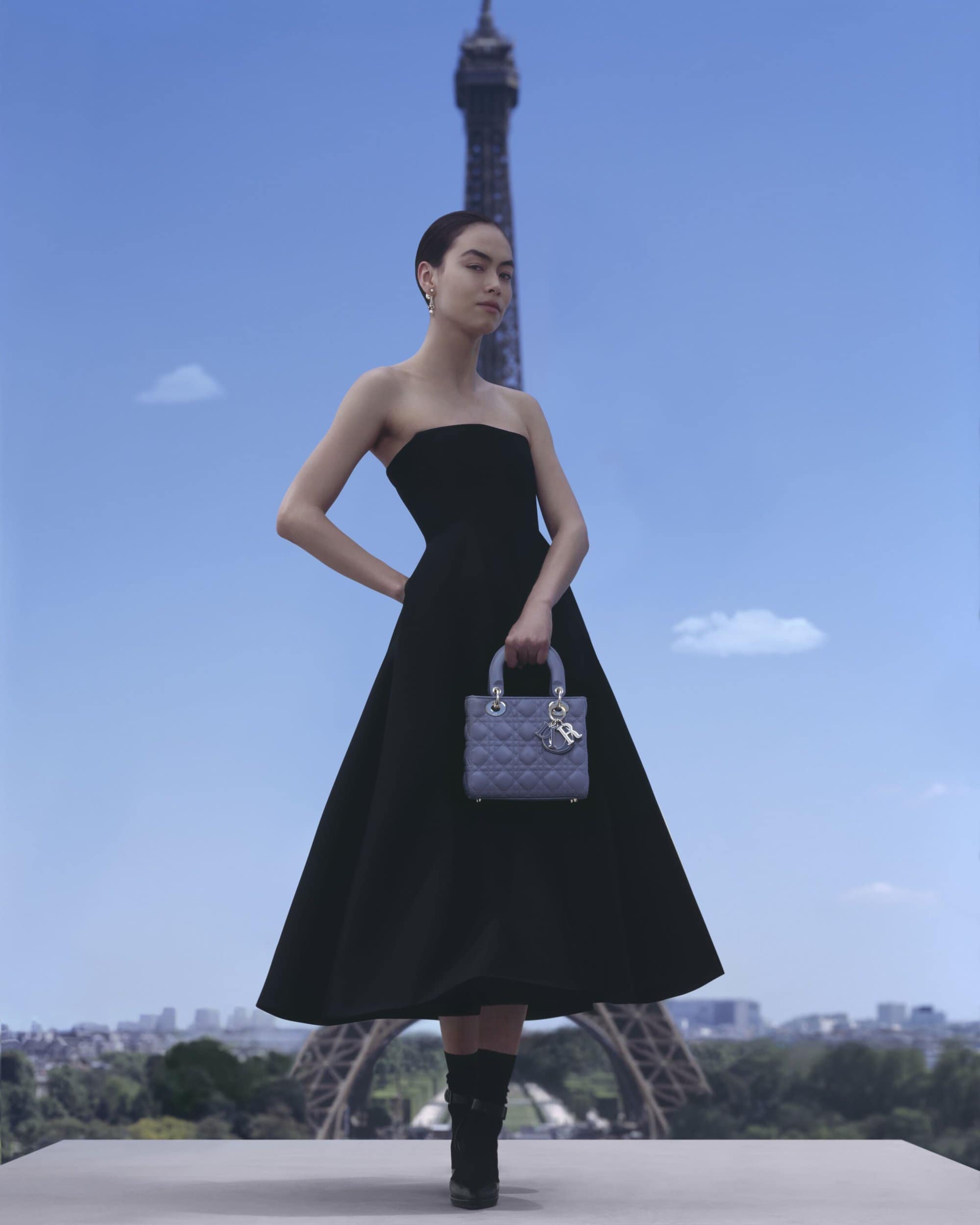 Woman in black dress holds handbag, Eiffel Tower in background, city skyline. Fashion editorial, outdoor, clear sky, urban setting.