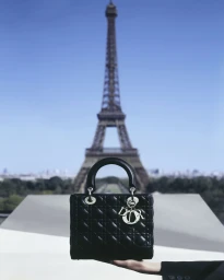 A black quilted Lady Dior handbag held aloft in front of the Eiffel Tower against a clear blue sky in Paris.