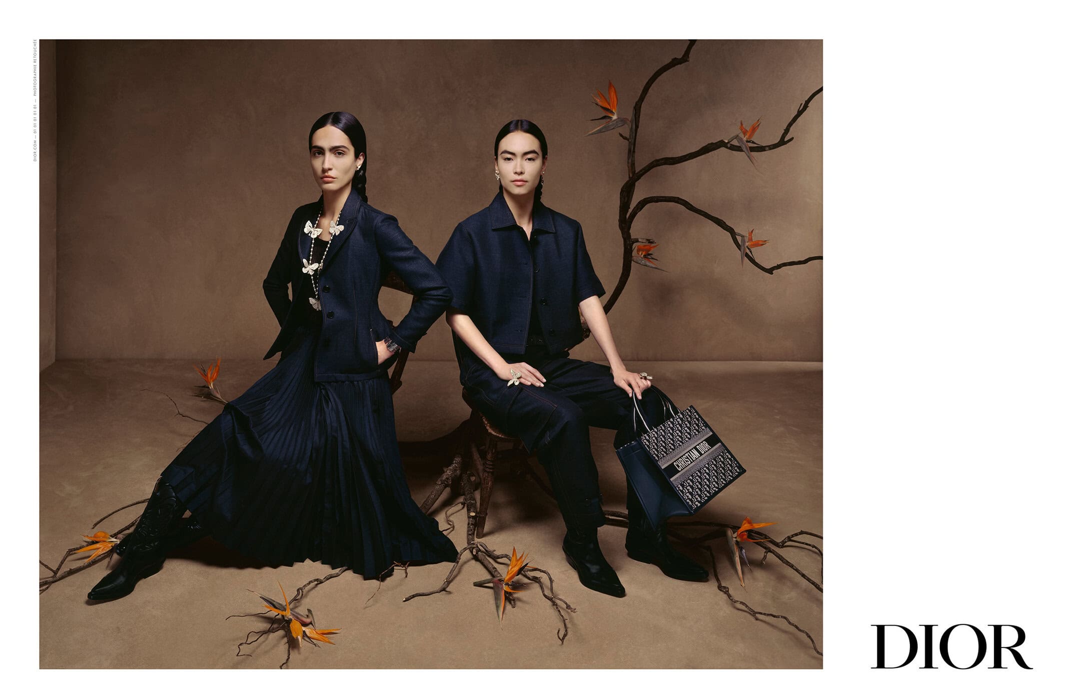 Two women model Dior apparel and accessories against a neutral backdrop with decorative branches and bird of paradise flowers.