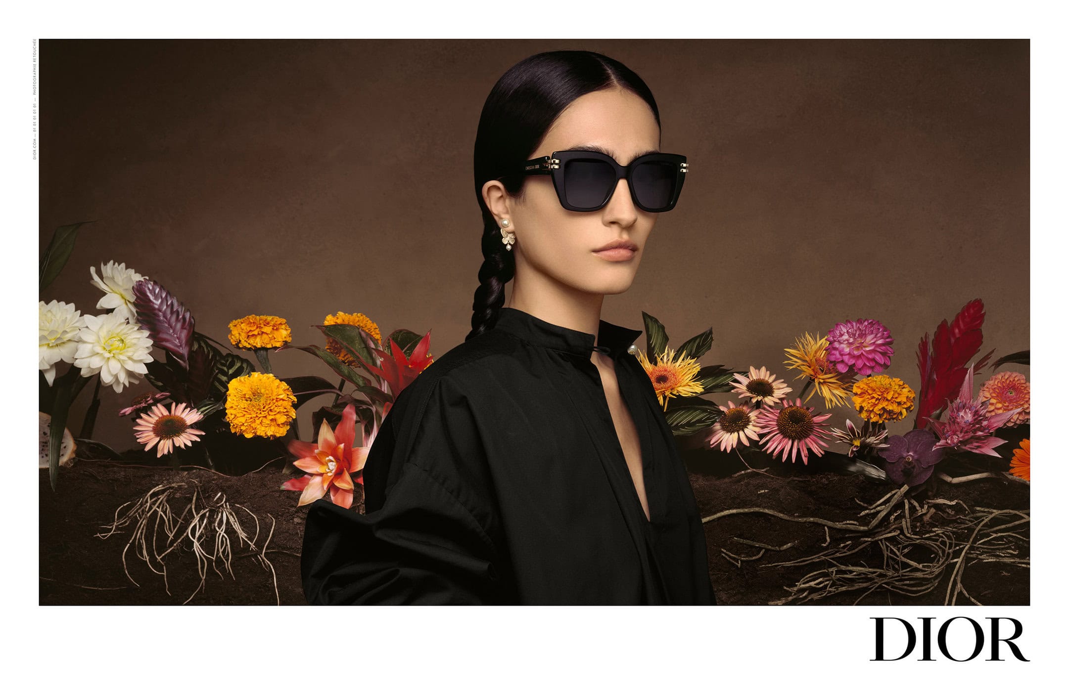 A woman wears black square sunglasses and a black top, posing in front of colorful flowers and a brown background, fashion editorial.
