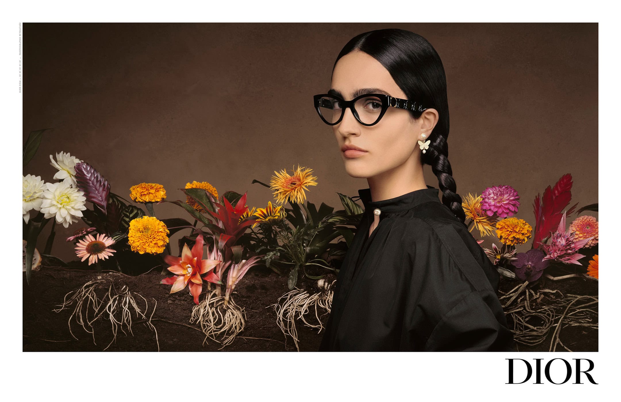 Elegant woman with dark hair in braids wears glasses and a black shirt against a backdrop of colorful flowers and exposed roots.