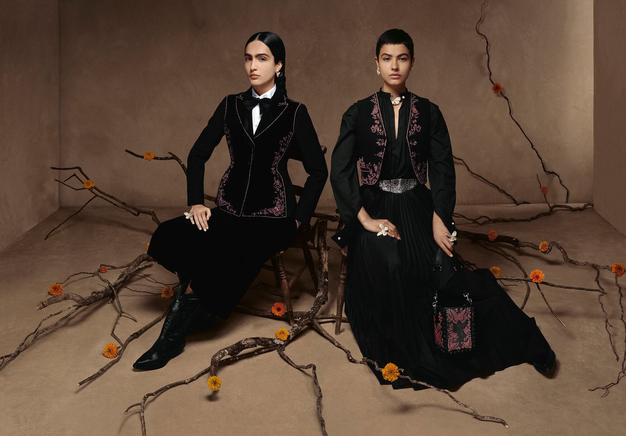 Two women in black velvet jackets and long skirts sit among branches with small yellow flowers in a studio setting, fashion shot.