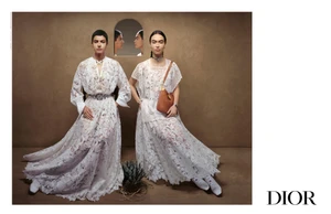 Two models in floor-length white lace dresses seated against a textured beige backdrop with an arched mirror and small agave plant.