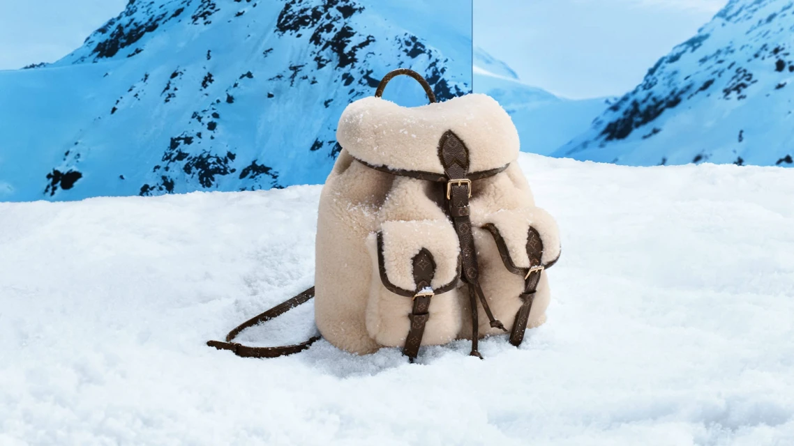 Fur backpack with leather straps and accents sits in the snow with snow-covered mountains in the blurred background on an overcast day.