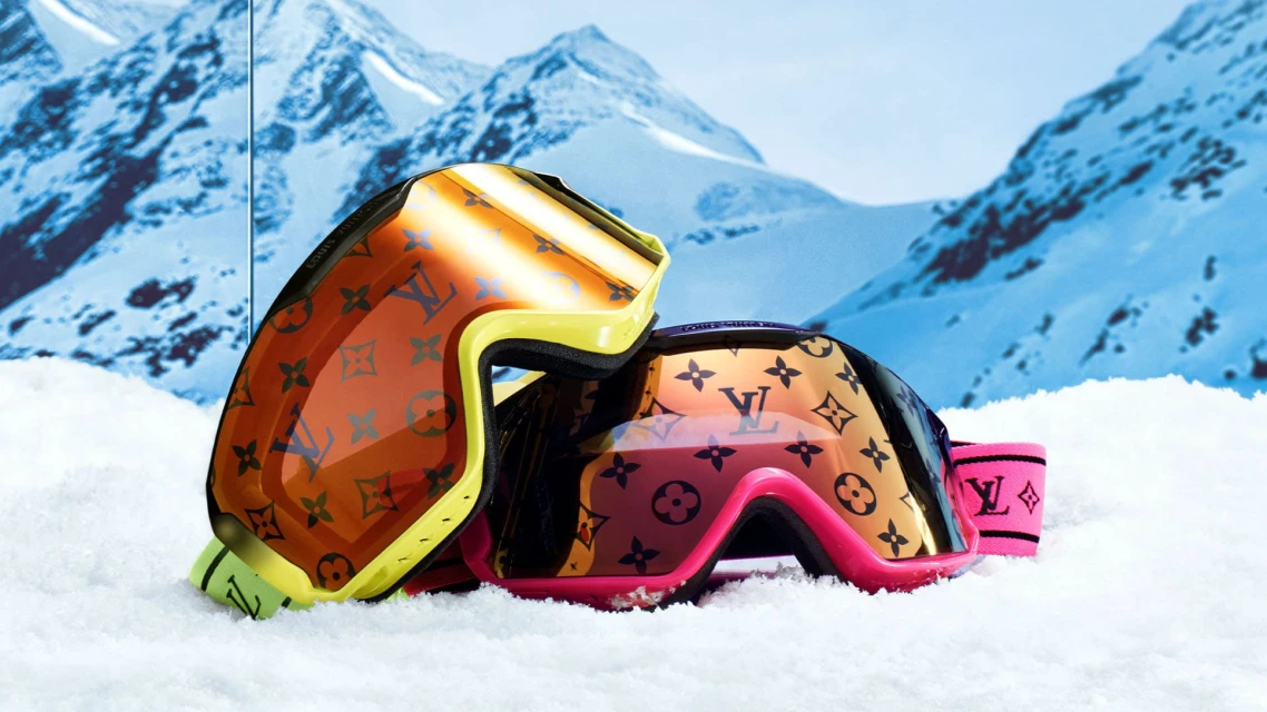Close-up studio shot of luxury designer ski goggles with the Louis Vuitton monogram on snow, with a snowy mountain range backdrop.