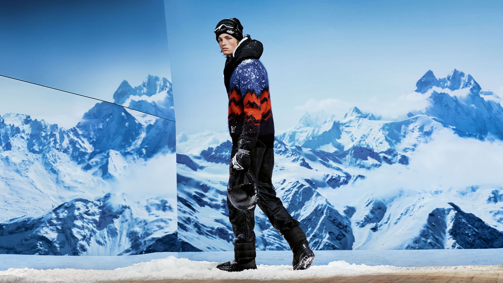 Male model in a patterned ski sweater and black snow pants standing against a snowy mountain backdrop