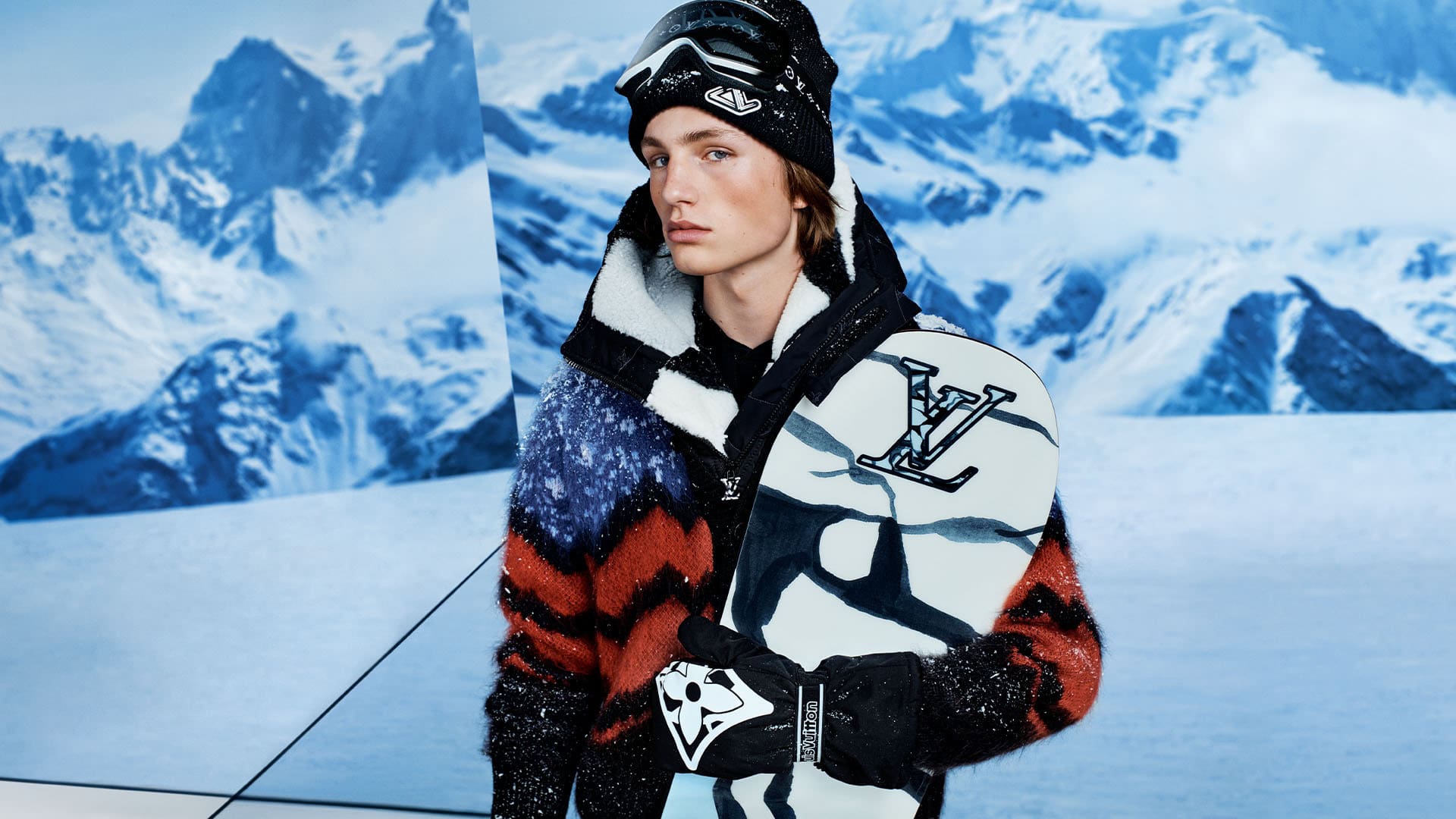 Male fashion model wearing a wool jacket, hat, and ski goggles holding a snowboard in a snowy mountain landscape.