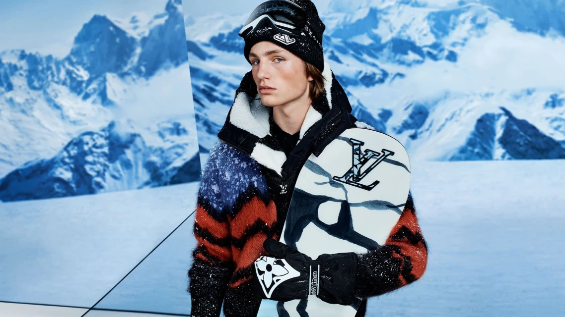 Male fashion model wearing a wool jacket, hat, and ski goggles holding a snowboard in a snowy mountain landscape.