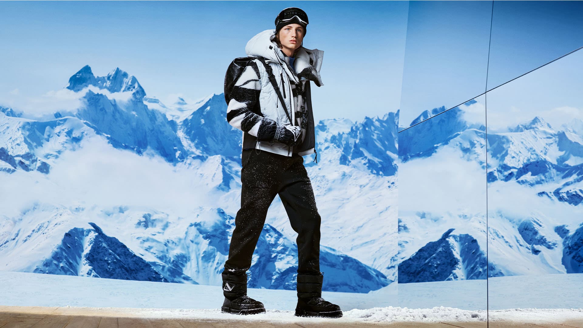 A fashionable adult stands in snowy mountains wearing ski goggles, black snow pants, and a heavy winter coat with mountain reflections.