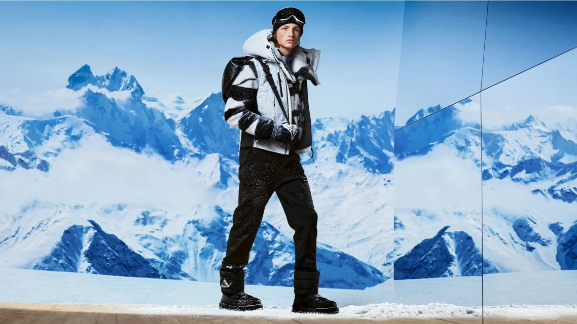 A fashionable adult stands in snowy mountains wearing ski goggles, black snow pants, and a heavy winter coat with mountain reflections.