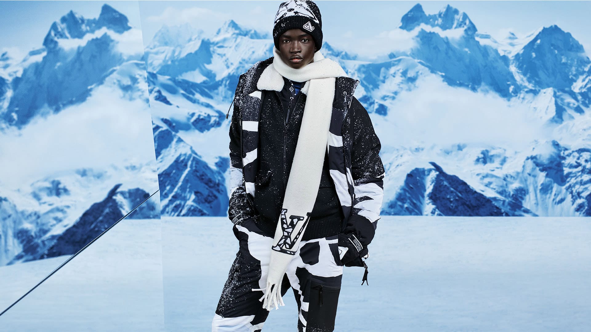 Model in a patterned ski suit, hat, and scarf in front of a snowy mountain range, showcasing winter fashion and outdoor adventure style.