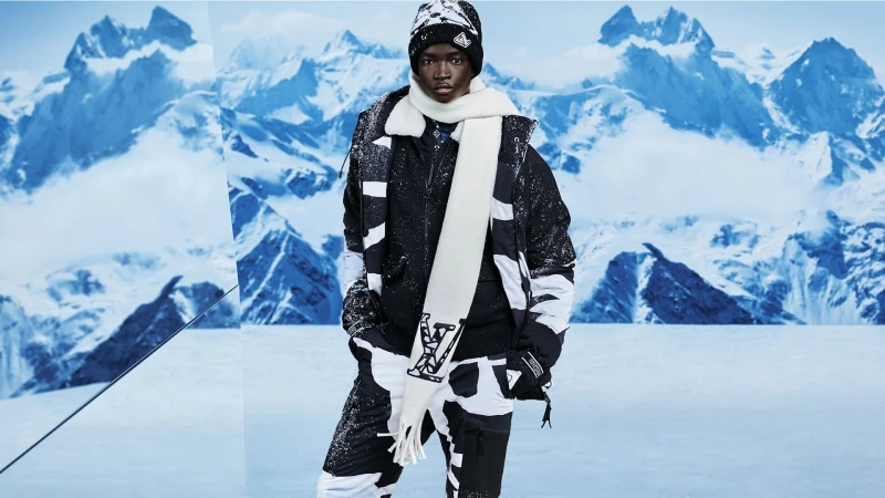 Model in a patterned ski suit, hat, and scarf in front of a snowy mountain range, showcasing winter fashion and outdoor adventure style.