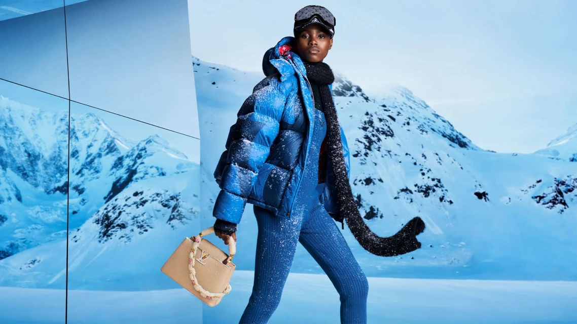 Fashionable woman in snow gear with handbag against a snowy mountain backdrop, posing in matching blue puffer jacket and jumpsuit.