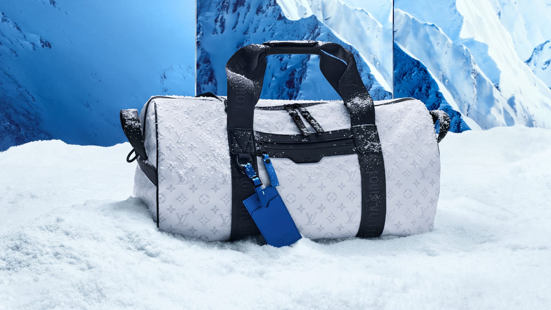 White Louis Vuitton duffel bag with black straps rests in the snow against a winter mountain background, luxury travel aesthetic.