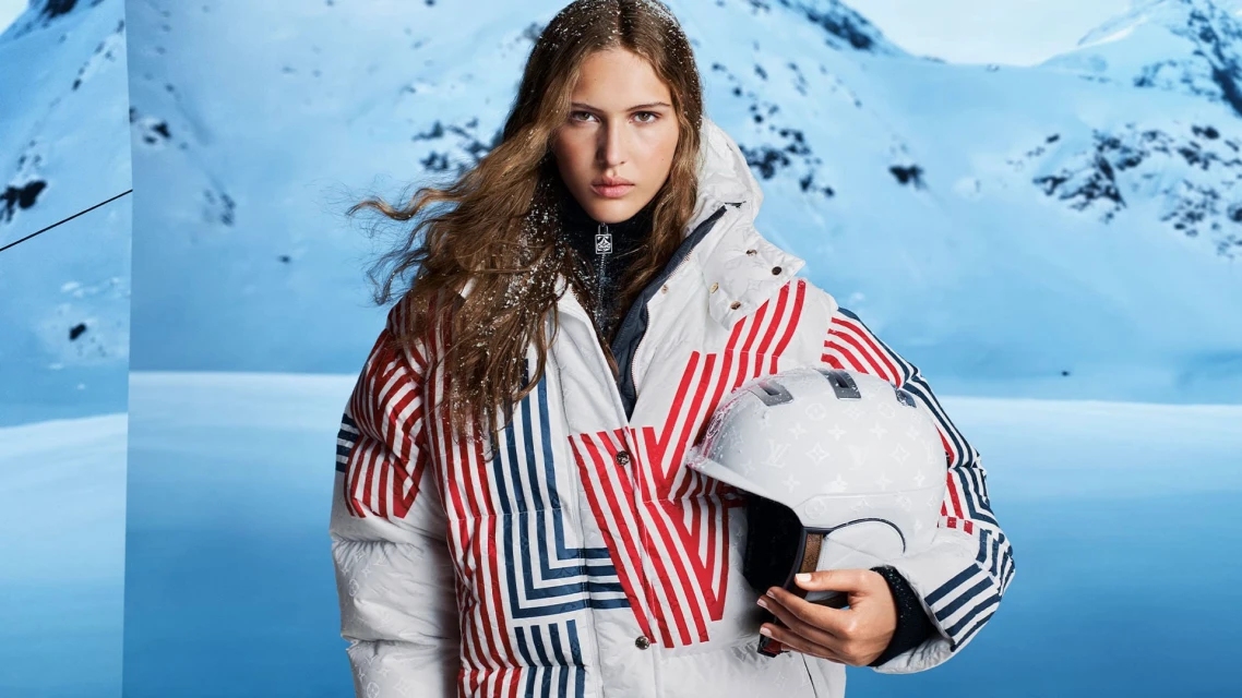 A woman in winter clothing poses outdoors with a helmet against a snowy mountain backdrop, creating a seasonal fashion image.