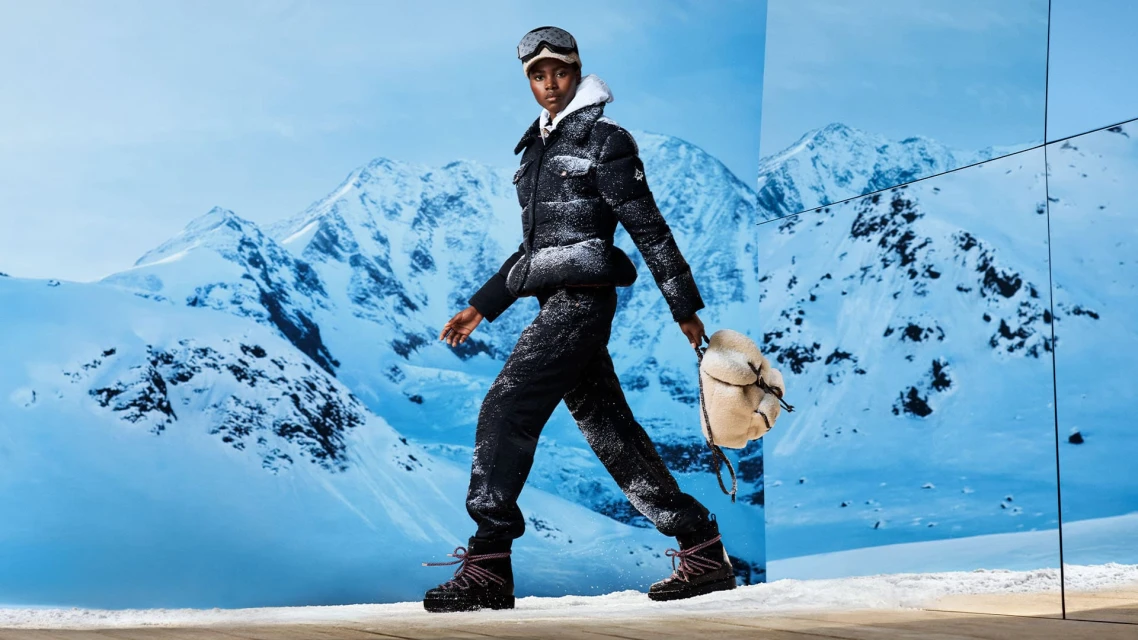 Woman in winter fashion walking against snowy mountain backdrop wearing puffer jacket, snow pants, boots, goggles, and carrying backpack.