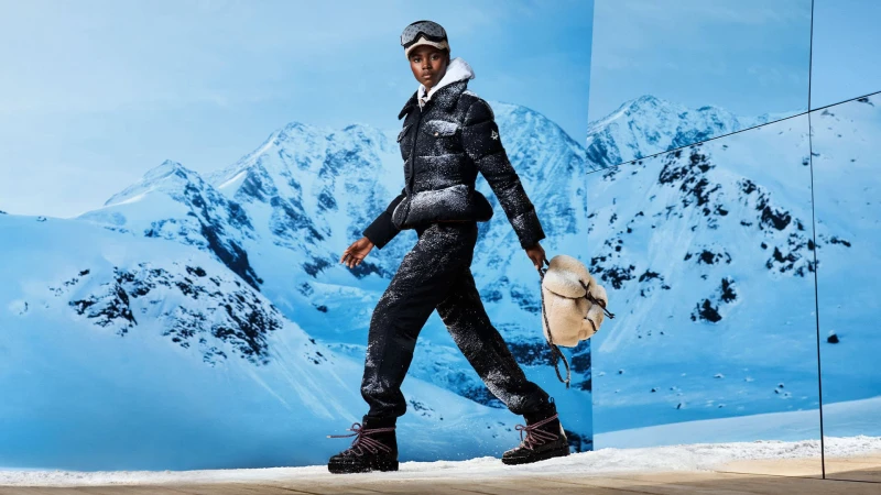 Woman in winter fashion walking against snowy mountain backdrop wearing puffer jacket, snow pants, boots, goggles, and carrying backpack.