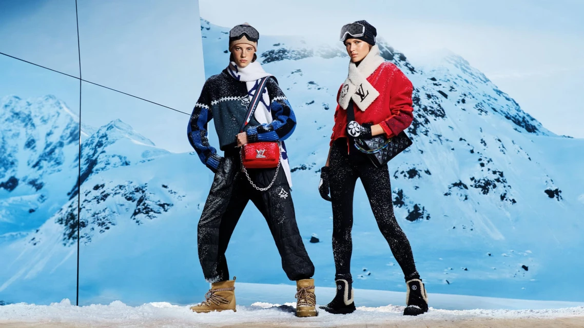 Two fashion models pose in ski apparel with mountains as a backdrop, showcasing winter style with hats, goggles, bags, and snow boots.
