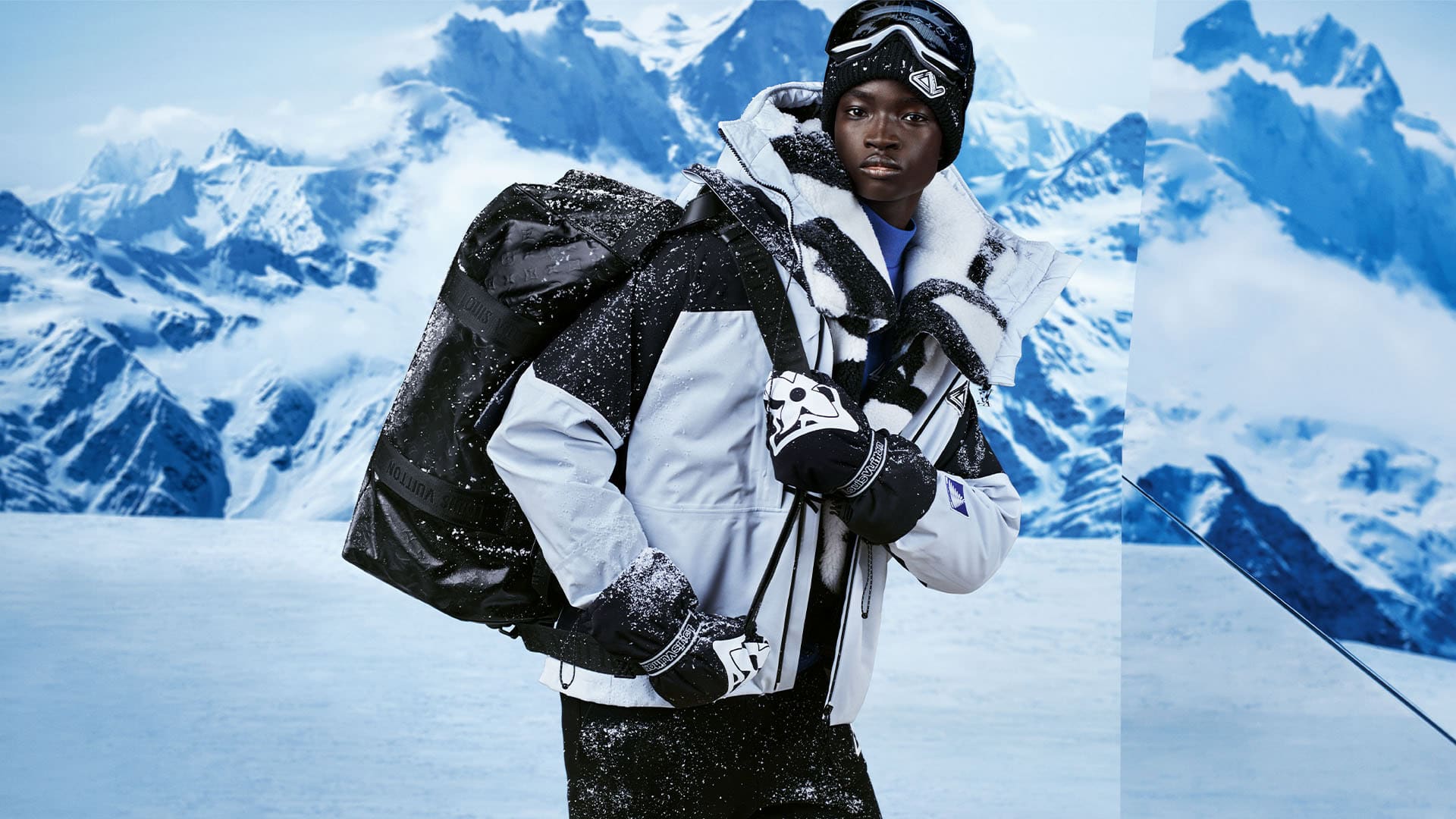 Man poses wearing ski goggles, beanie, black and grey jacket, gloves, and carries a duffel bag in a snowy mountain setting.