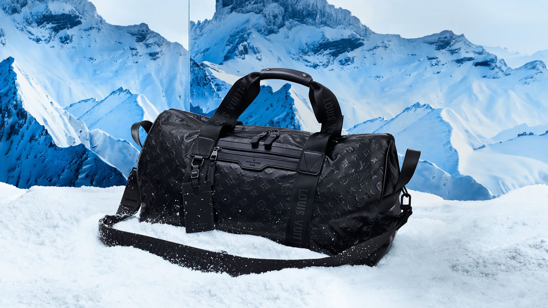 A black Louis Vuitton duffel bag sits in snow against a backdrop of snowy mountains, highlighting luxury travel gear in a winter setting.