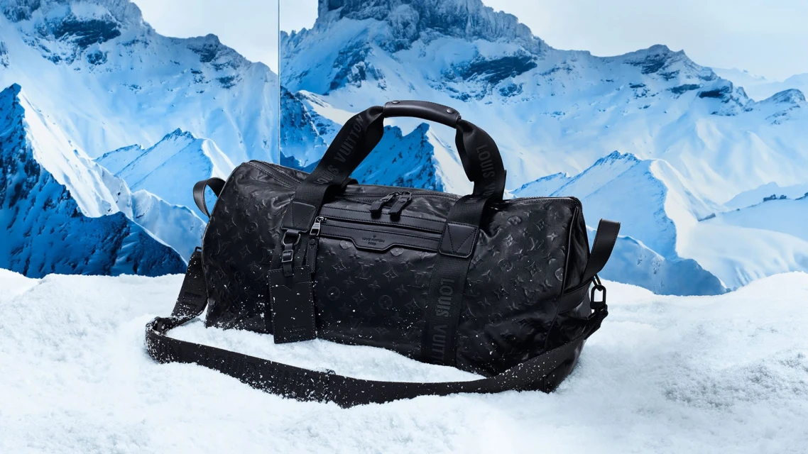 A black Louis Vuitton duffel bag sits in snow against a backdrop of snowy mountains, highlighting luxury travel gear in a winter setting.