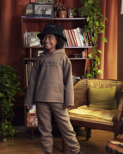 Portrait of a smiling child in a brown hoodie and matching sweatpants, wearing a black bucket hat in a cozy, vintage-style interior.