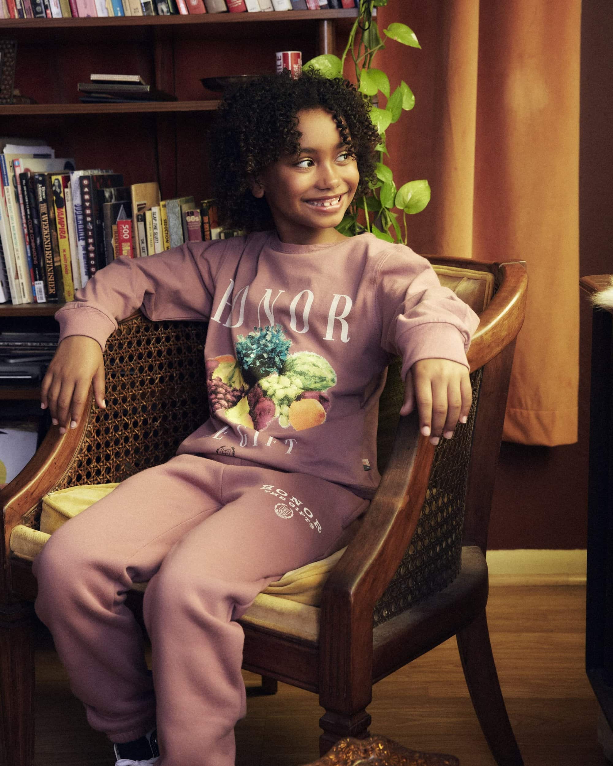 Child in a mauve sweat suit with a fruit graphic, sitting in a wicker chair in front of a bookshelf, looking away and smiling slightly.