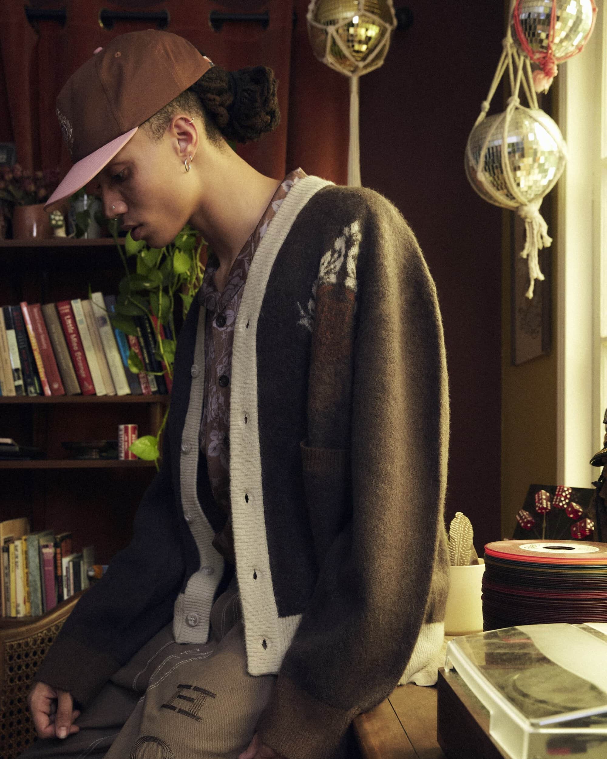 Person wearing a brown baseball cap and cardigan sweater posing indoors near a bookshelf and record player, bathed in soft light.